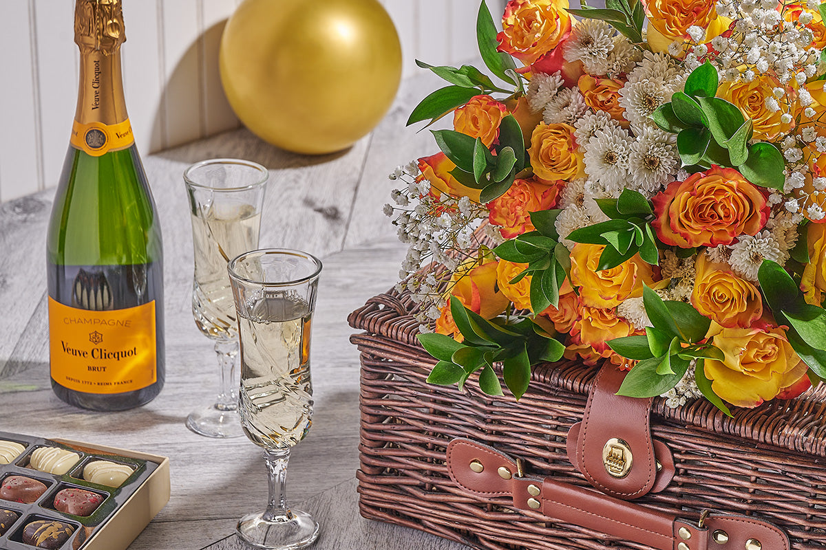 Bottle of Veuve Clicquot champagne, glasses with champagne, chocolate truffles, and a floral basket on a wooden table.